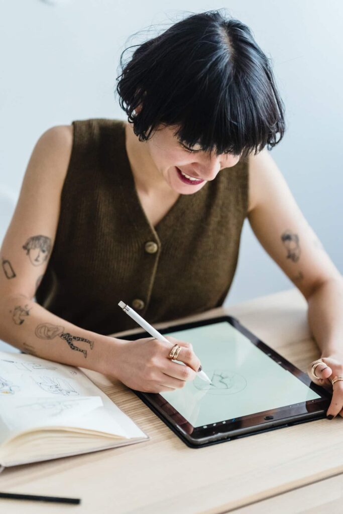 smiling-woman-drawing-on-graphic-tablet-with-pencil-at-table - howtospotify 